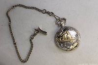 Metal pocket watch with embossed steam locomotive and chain displayed on white background.
