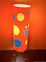 Tall orange cylindrical table lamp with colorful polka dots photographed against an orange background on a wooden surface. Lamp has thin metal legs with ball feet.