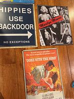 Photo showing all three metal signs arranged on wooden surface: 'Hippies Use Backdoor', Marilyn Monroe collage, and 'Gone with the Wind' movie poster sign.