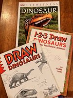 Set of three dinosaur books shown fanned out on table: DK Eyewitness Dinosaur, Draw Dinosaurs, and 1-2-3 Draw Dinosaurs and Other Prehistoric Animals by Freddie Levin.