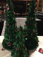 Three green foil-covered metal frame Christmas trees decorated with red beads displayed on a white surface with a living room background.