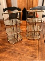 Two gold framed mini shelves with glass shelving and mirrored backs shown from the front angle on a wooden table.