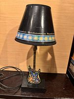 Desk lamp shaped like a colorful Stratocaster guitar with a black lampshade featuring decorative sun pattern and 'Masterblasterstratocaster' text