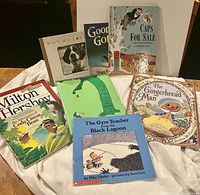 Photo showing seven children's books arranged on a white cloth, titles visible including Milton Hershey Cocoa Bean story, The Giving Tree, The Gingerbread Man, and others.