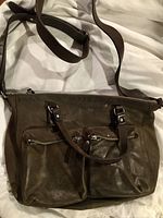 Brown leather messenger bag with two front zippered pockets, double handles, and adjustable shoulder strap laid out showing front and back