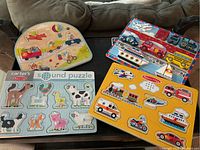 Four children’s puzzles arranged on black table, showing colorful vehicles, animals, and circus themes. Some puzzles include peg handles for easy grasping.