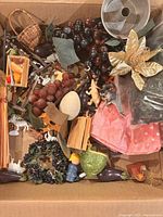 Photo showing box containing assorted miniature plastic grapes, wooden and plastic animals, small toys, miniature furniture pieces, fabric flower, and other craft items in various colors.