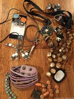 Photo shows assortment of rings, bracelets, necklaces and earrings spread on wooden surface, including multi-wrap purple leather rhinestone bracelet, turquoise beaded bracelet, peach bead necklace, black stone pendant necklace, and assorted metal rings and earrings.