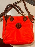 Front and top view of bright orange oversized faux leather bag with brown leather straps and gold emblem on front.