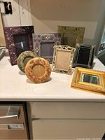 Overview image showing all 7 frames arranged on a white surface, including fabric and beaded frames, a gold mirrored frame, and a white metal frame