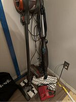Two Shark vacuums standing against wall with cords loosely coiled, various attachments and a red charger visible beside them.