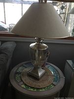 Full view of the table lamp on a circular patterned table with beige conical shade and silver finish base.