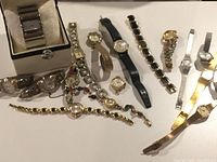 Lot of 13 watches arranged on a white surface revealing variety of bands and styles including bracelet style and stretchy metal links.