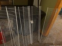 Four white wire closet shelves standing vertically, leaning against a dark background, showing their grid pattern and white coated metal rods.
