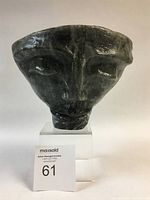 Front view of dark textured cast glass face sculpture on clear stand.