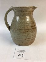 Jug front facing showing round body, handle on left, and spout on right with visible glaze texture and brownish banding.