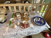 Photo of various assorted china pieces arranged on a lace tablecloth, including cups, saucers, salt and pepper shakers, a blue and white divided plate, and a glass pitcher.