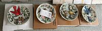 Four Rhodes Studios collector plates laid out on boxes. Each plate has distinct songbird and floral sculpted designs.