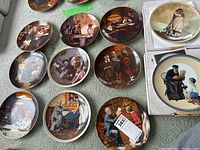 Overview photo of 11 Knowles collector plates laid out on surface showing various scenes including people in indoor settings, domestic and social themes.