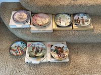 Seven Asian collector plates displayed on stairs, some on white cardboard boxes, various cultural and floral themes.
