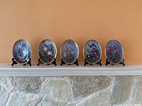 Front view of five oval collector plates on stands showing floral artwork and gold rims.