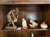 Four decorative pieces displayed on wooden shelving including a carved wooden figure and three smaller figurines.