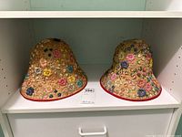 Two beige bucket hats with dense multicolored floral embroidery and red rim trim displayed on a white shelf.