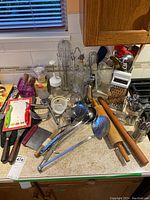Overview of assorted kitchen utensils including spatulas, ladles, rolling pin, metal grater, tongs, bottles and other tools on countertop.