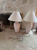 Two matching table lamps with beige ceramic ribbed bases and white fabric shades, placed on carpet in front of stone fireplace.