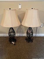 Two black metal base lamps with beige fabric shades on carpeted floor against wall, showing entire lamps