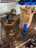 Overview photo of lot items on workbench: two vintage oil lamps, Coleman Sport Light Lantern box, and blue flashlight