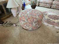 Round floral upholstered ottoman positioned on carpeted floor near lamps and floral patterned couch.