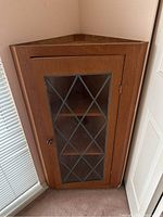 Wooden corner cabinet with glass door, locked in corner space against wall and carpeted floor.