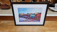 Full view of the framed signed urban landscape print on a wooden surface.