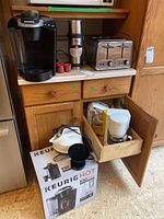 Photo showing Keurig K50 coffee maker on box, stainless steel toaster, Thermos kettle, coffee pods on a kitchen counter beside a small cabinet with drawer open revealing more appliances.