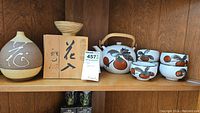 Photo of wooden shelf with Japanese ceramic tea pot decorated with orange fruit motifs, five matching tea cups, ceramic vase, wooden bowl, and wooden box.