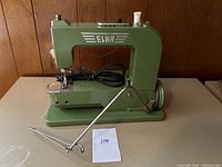 Full side view of vintage green Elna sewing machine with metal sewing guide accessory in front and black power cord.
