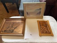 Lot of three framed wall art pieces, placed on a table, including a framed mountain print, a wooden ship carving, and a smaller wooden relief carving.