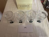 Four Ralph Lauren heavy scotch glasses arranged side by side with a Lauren Ralph Lauren box in the background and a small card with number '59' in the foreground.