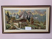 Framed needlepoint artwork showing complete landscape with mountains, cabin, watermill, trees, and sky.