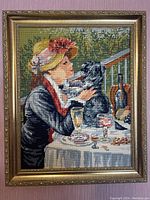 Full view of framed needlepoint mounted on purple wall showing woman with hat holding dog at table with bottles and glasses.