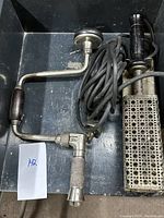 Photo showing both the hand crank drill with wooden handles and the electric soldering iron with power cord and metal stand in a dark box.