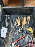 Top-down view inside open box showing assorted hand tools including wood chisels, pliers, files, and wire strippers.