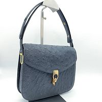 Front three-quarter view of bag on stand showing flap, texture and strap attachment.