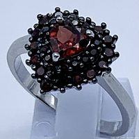 Close-up photo of heart-shaped center garnet stone surrounded by smaller garnet stones, all set in sterling silver with 14K white gold plating, showing rich red colors and detailed setting prongs.