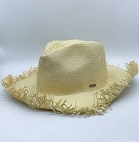 Front angled view of light beige straw fedora hat with frayed brim and metal plaque on crown side.