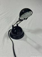 Front perspective showing the black dome-shaped lamp head, adjustable arm, round base, and attached power cord.