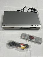 Top view of the silver Koss KS4112 DVD player with power cord visible, remote control and audio-video cables in plastic wrap placed in front.