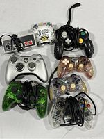 Eight game controllers including wired NES, Guitar Hero, Nintendo GameCube, Xbox 360, and translucent wired controllers arranged on a flat surface.