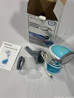 Handheld garment steamer set including steamer device with power cord, fabric brush attachment, water cup, user manual, and product box.